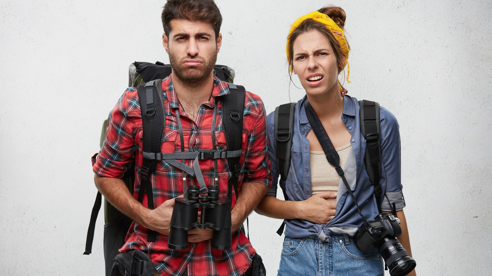 A man and a woman dressed like they're traveling with unhappy expressions.
