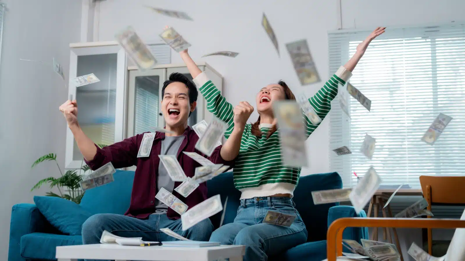 An excited couple sits on the couch throwing money in the air to represent financial success.