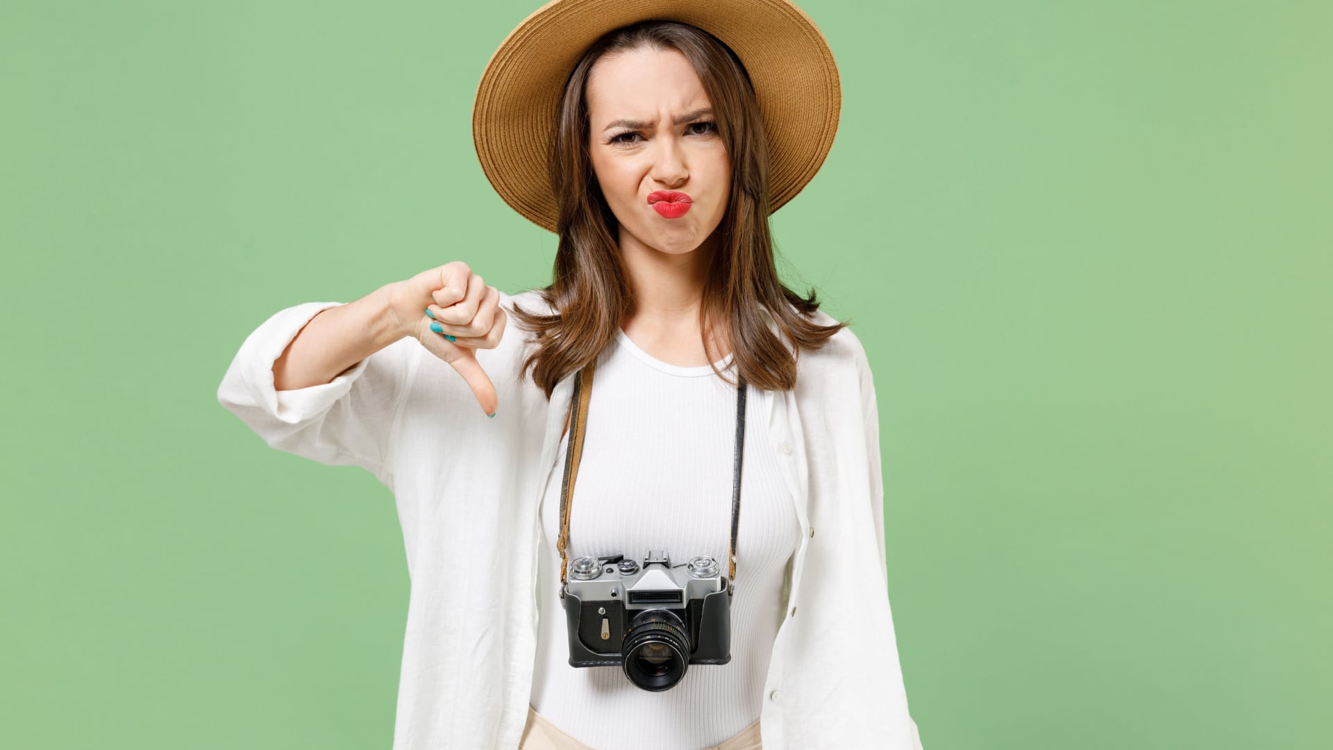 woman with travel hat and camera making an upset face and thumbs down sign on a green background.