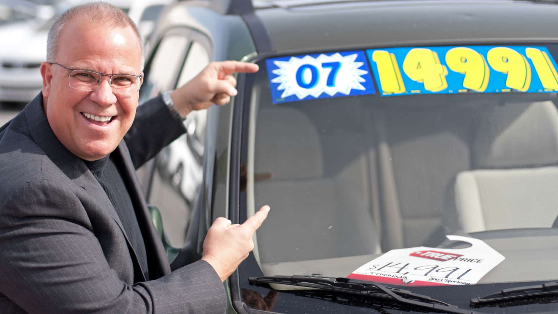 Car salesman on a lot pointing at the price on a car's windshield.