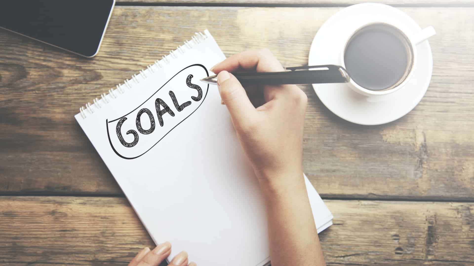 person writing the word "goals" on a notepad on a table next to a cup of coffee. 