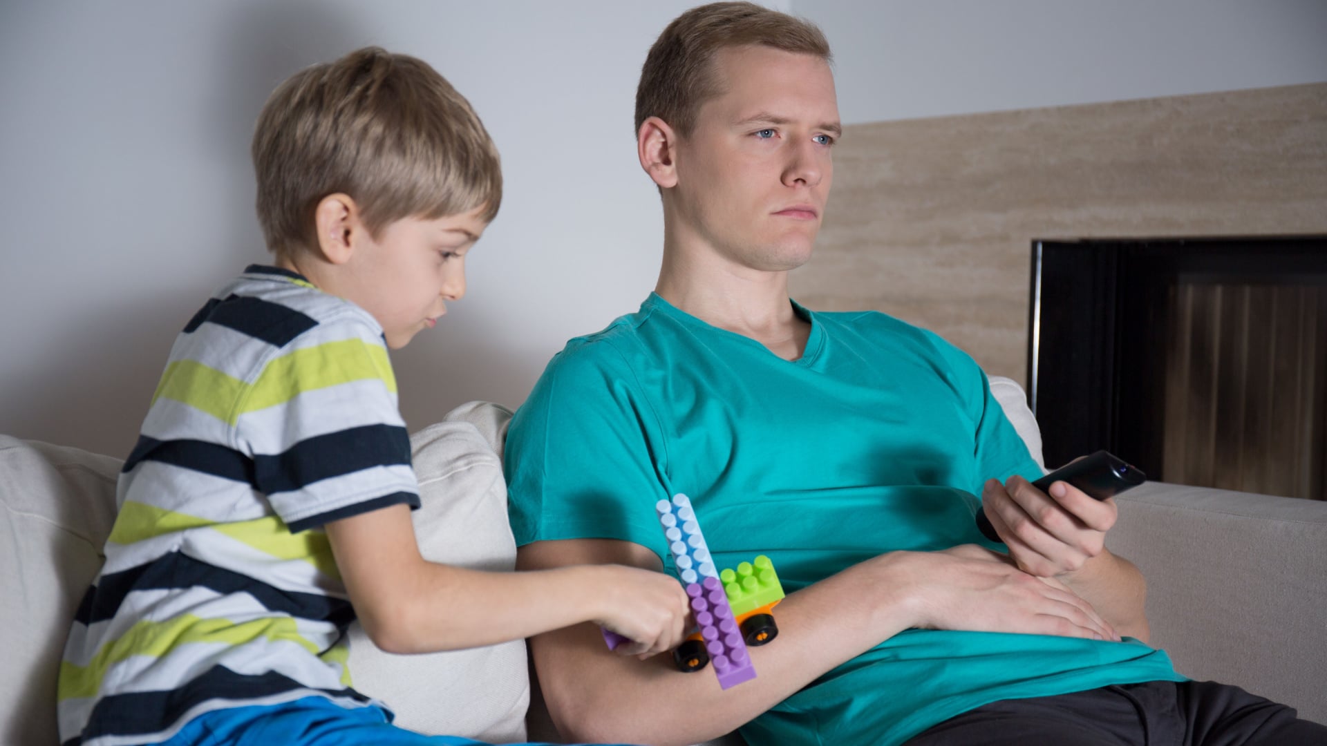 Dad ignoring his son and watching tv while son tries to play with a toy truck on dad's arm.