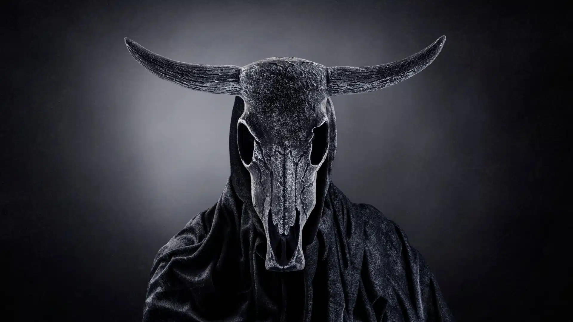 Black and white image of a scary monster. Cows skull with horns wearing a balck robe.