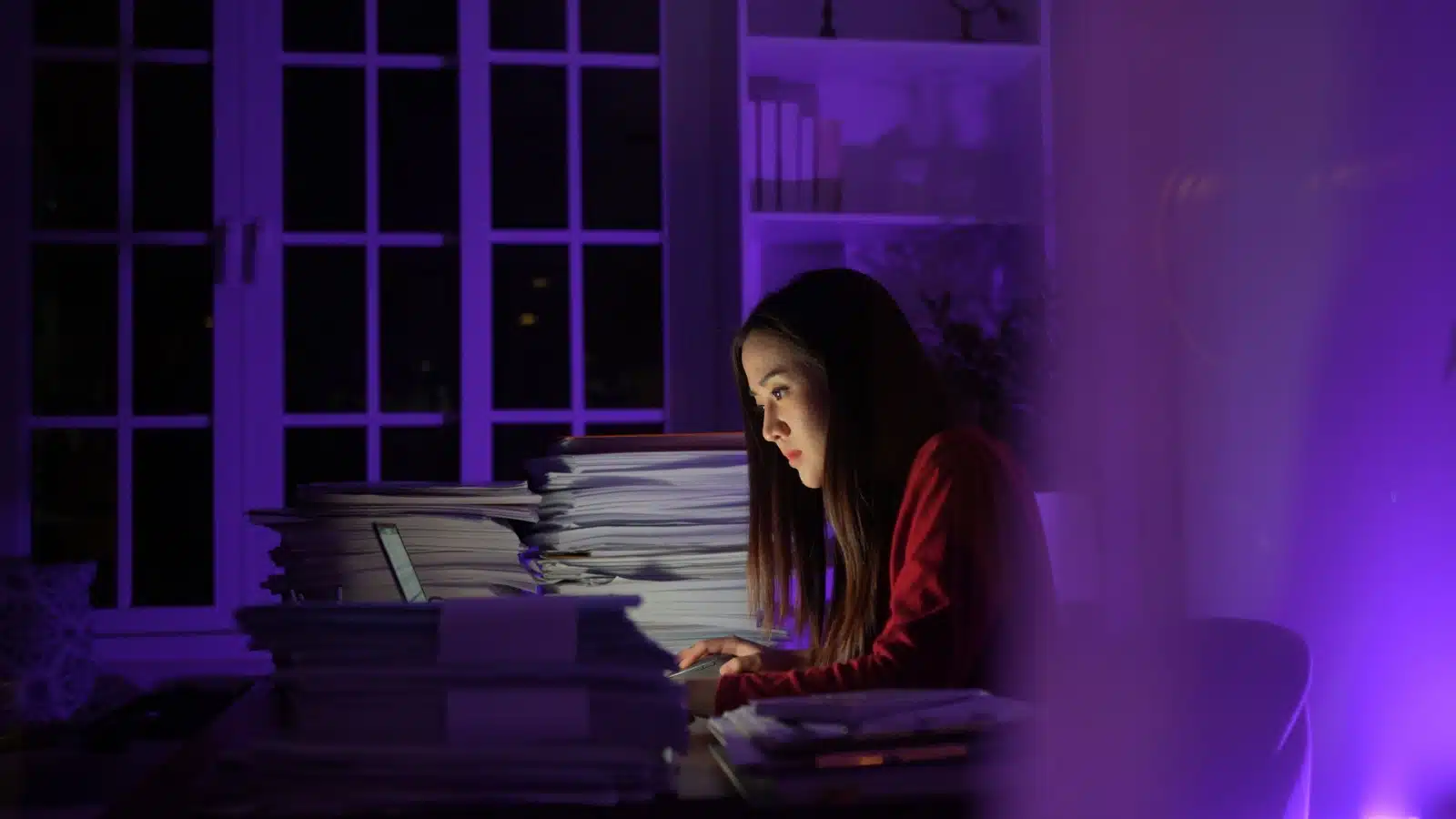 A woman works late into the night, surrounded by piles of more work.