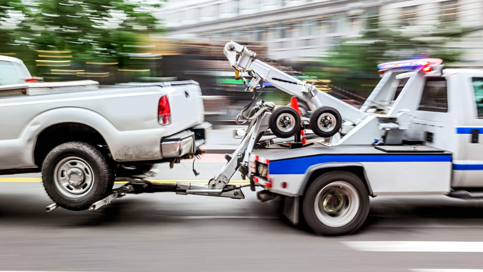 Small tow truck towing a pick up truck by it's rear side.