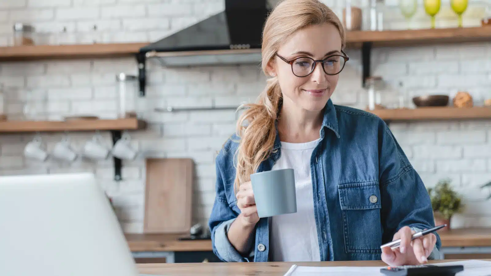 A happy woman sits in her kitchen calculating her bills to represent why you should pay yourself first.