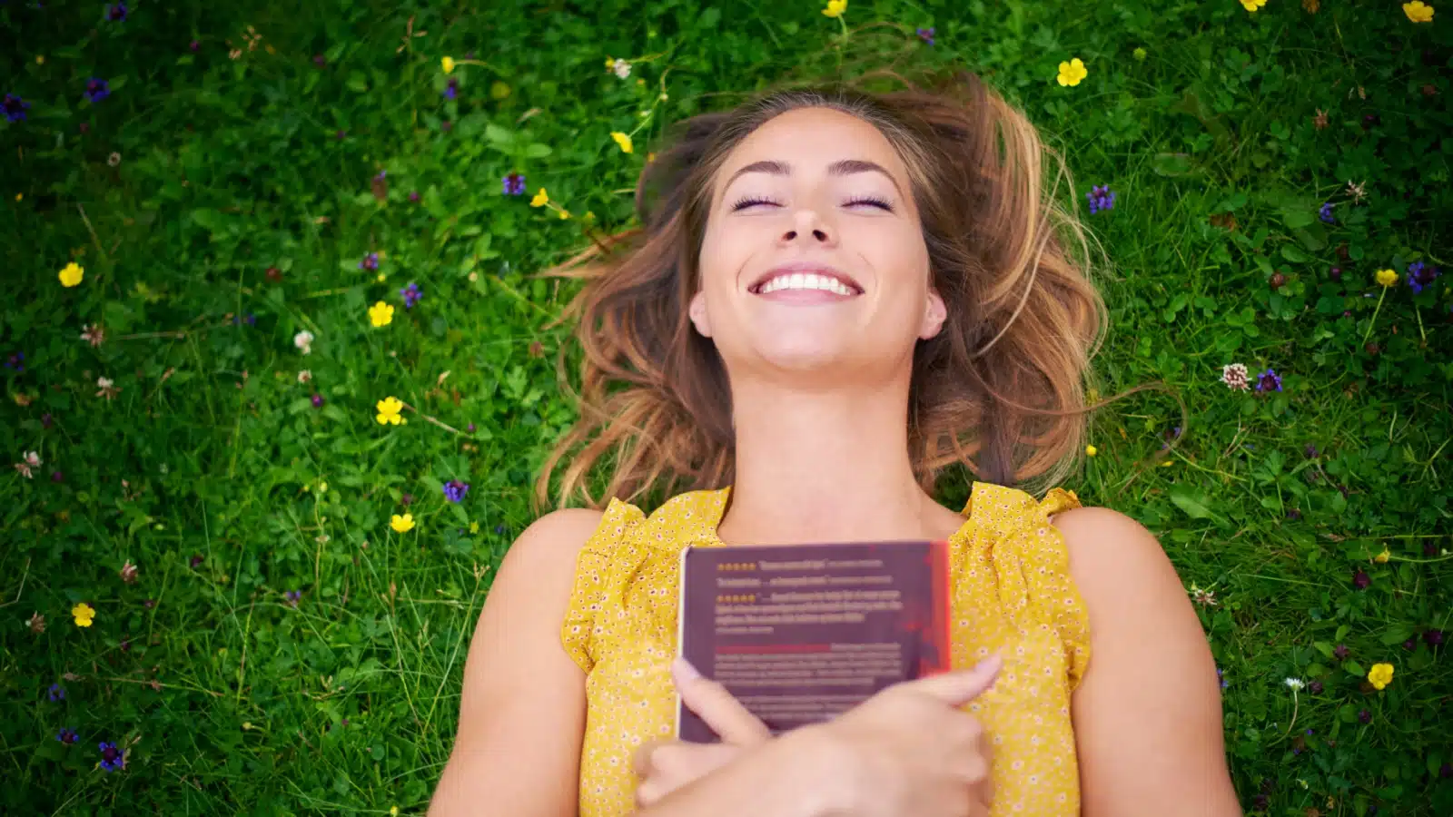 A woman lays in the grass, hugging her book after finishing it because it had a satisfying resolution.