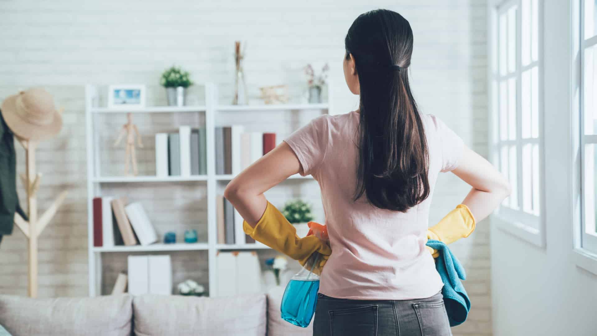 Woman looking away into a room with her back to the camera. She has her hands on her hips and cleaning supplies in them. She looks as if she's ready to tackle the chores.