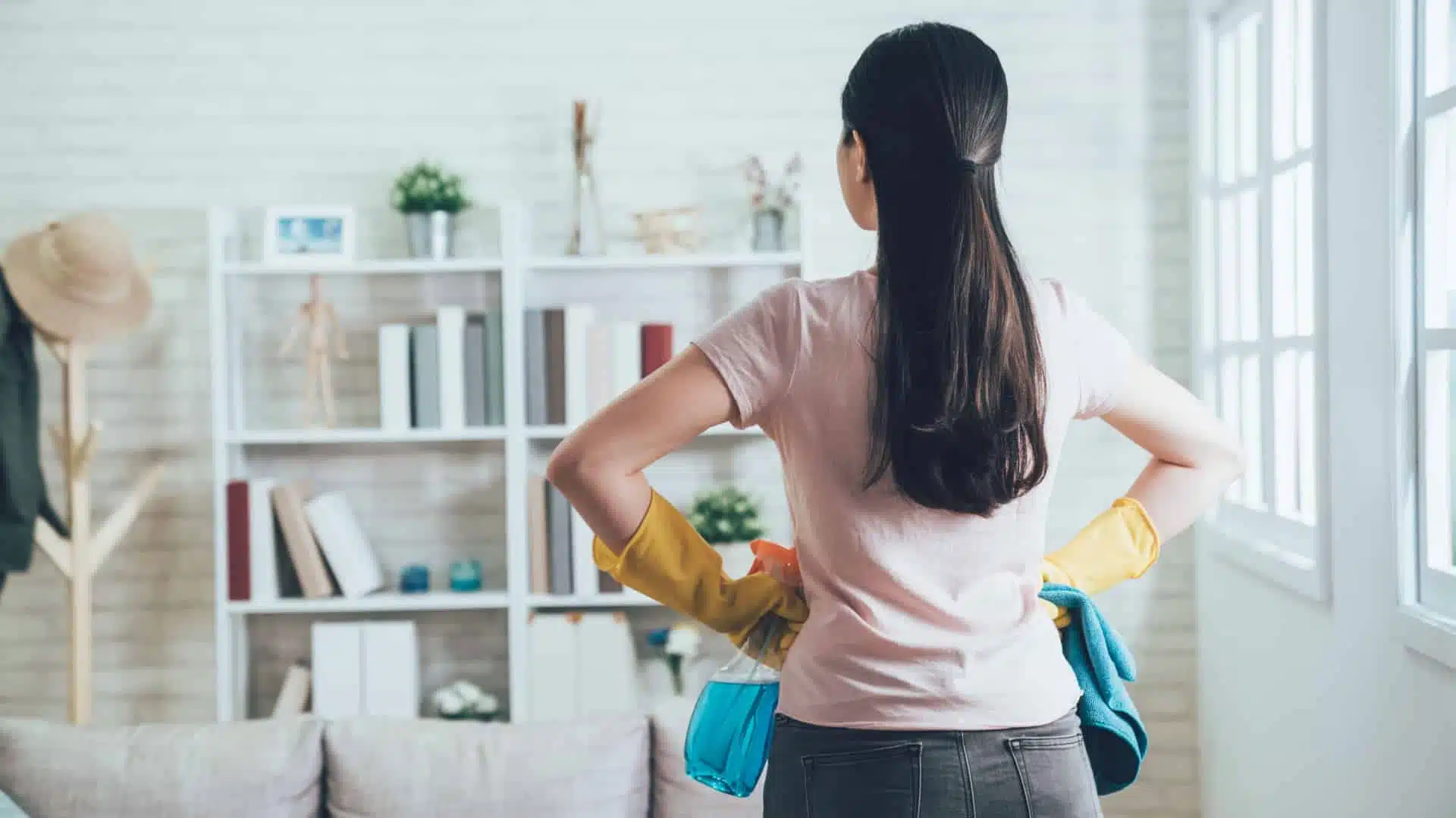 Woman looking away into a room with her back to the amera. She has her hands on her hips and cleaning supplies in them. She looks as if she's ready to tackle the chores.