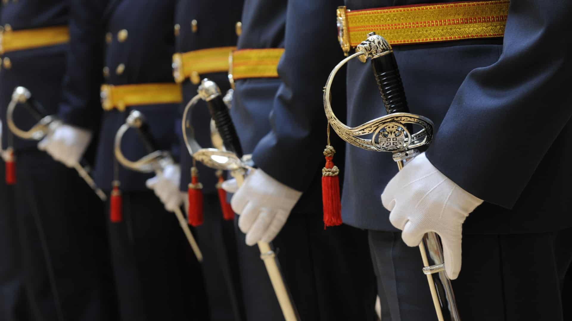 Military personnel dressed in fancy dress blues with their hands on their ceremonial swords.