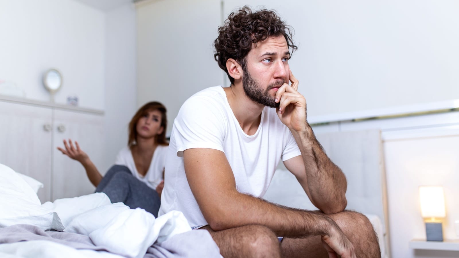 An upset man sulking in bed while his wife is in the background with her hands up.