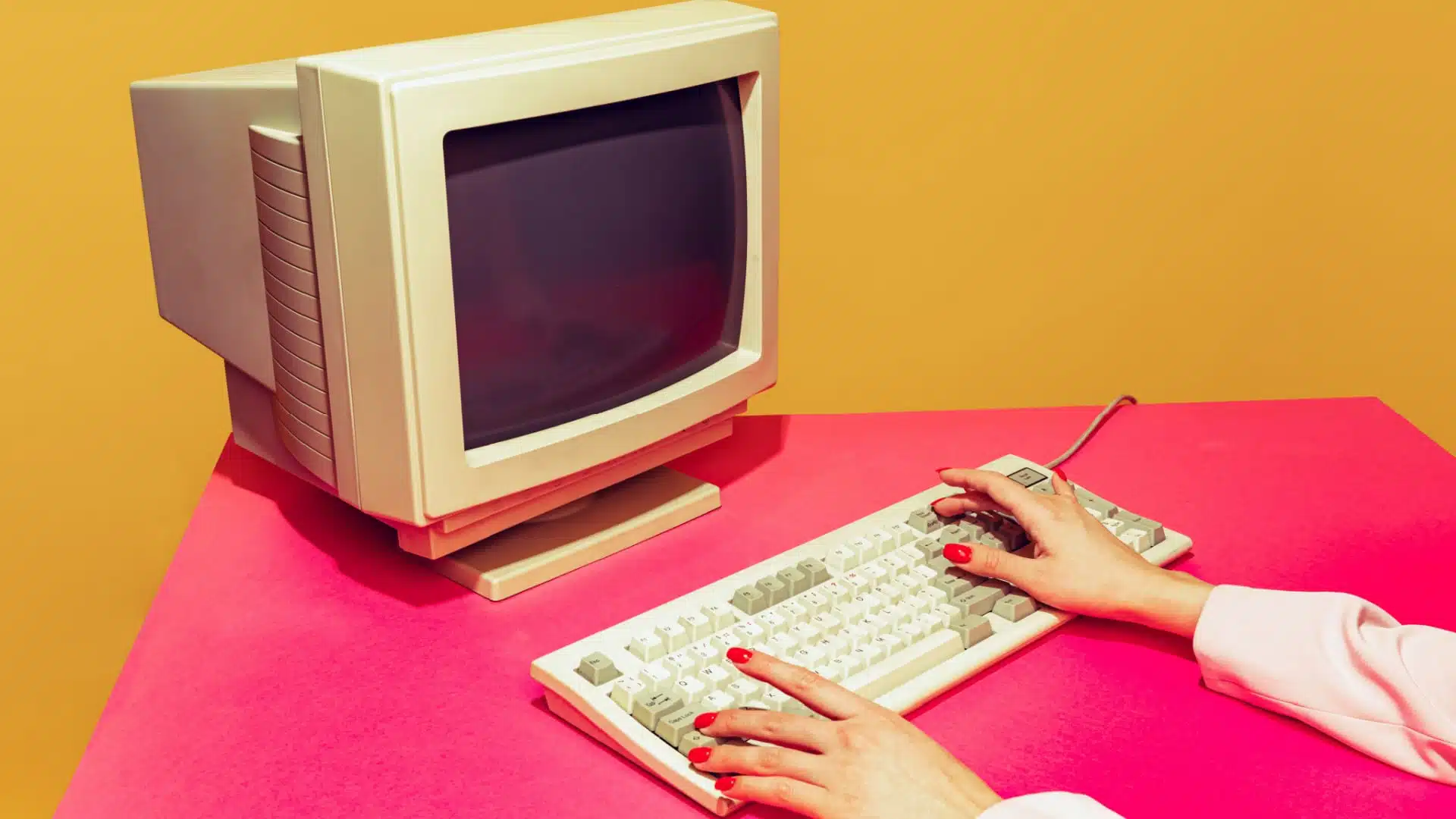 Vintage computer on a colorful bright pink desk against a bright orange wall.