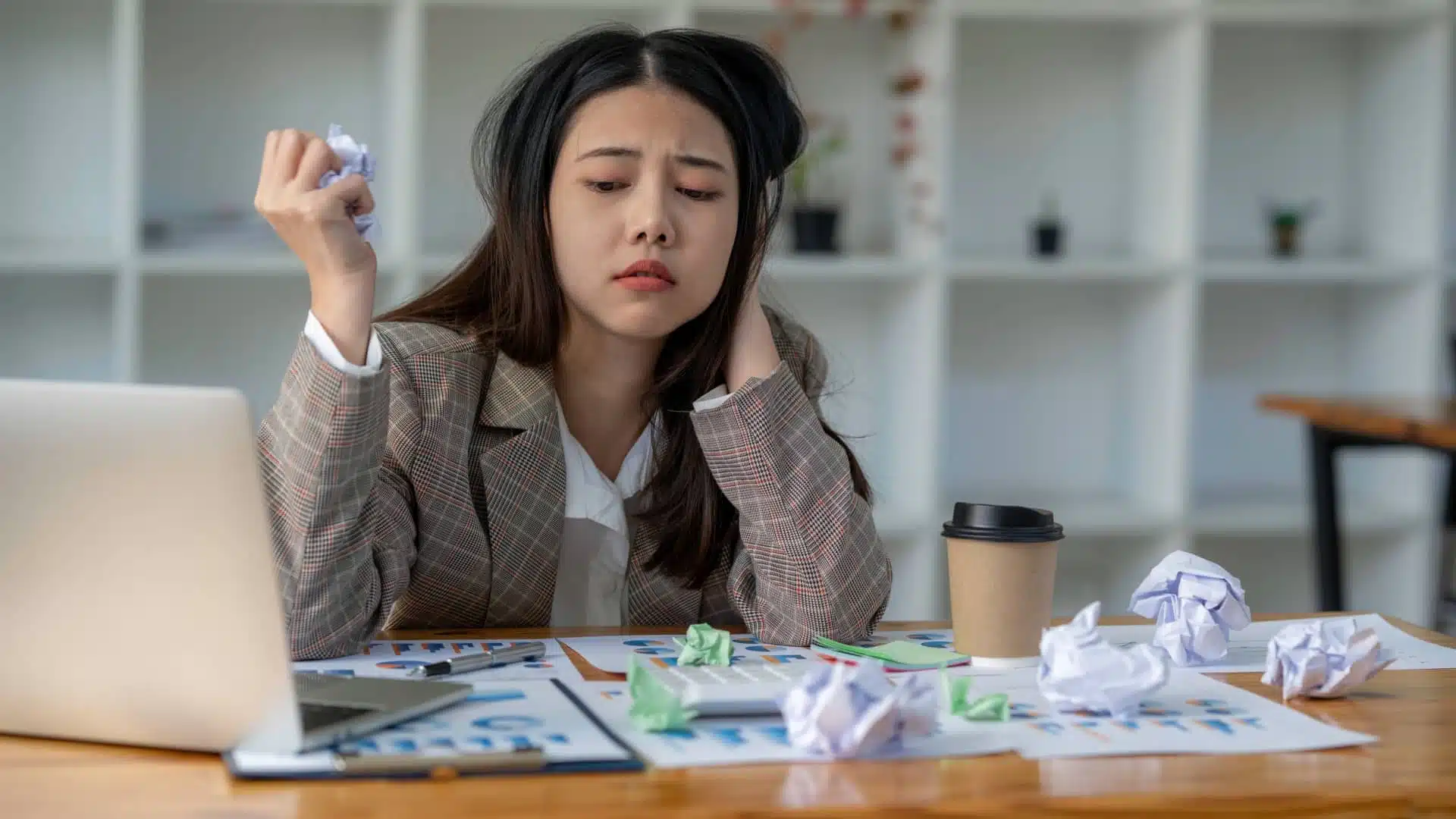 A defeated looking woman sits at a desk with a bunch of crumpled up papers on it. 