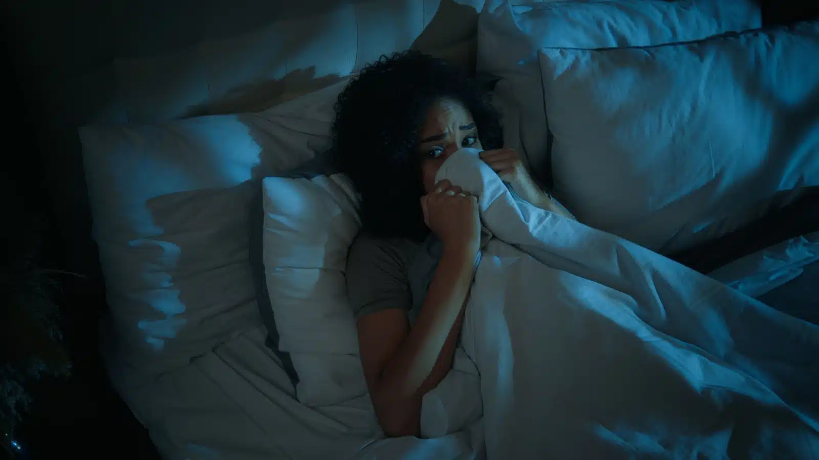 A woman lies in bed, pulling the covers over her face because she's terrified. She looks like she's seen or heard a ghost.