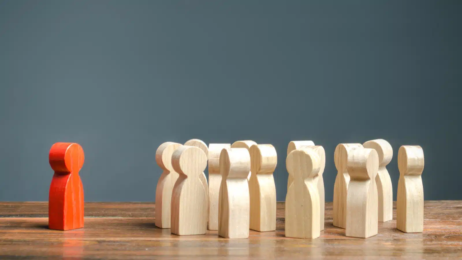 A red figurine stands away from a group of plain wooden figurines to represent an outsider or someone who is different.