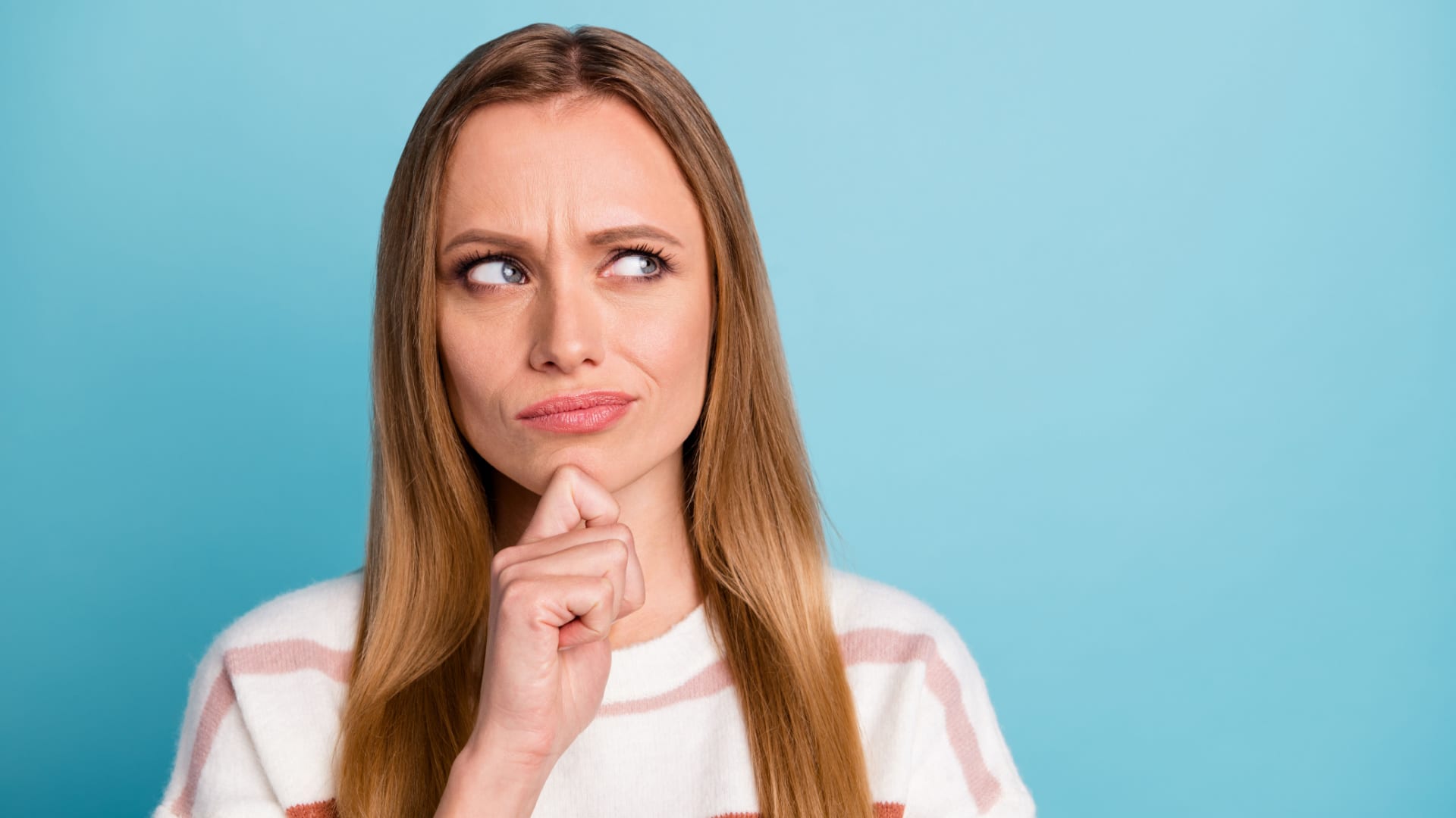 A woman looks unsure with her hand on her chin and her eyes looking up as if she's thinking about something serious.