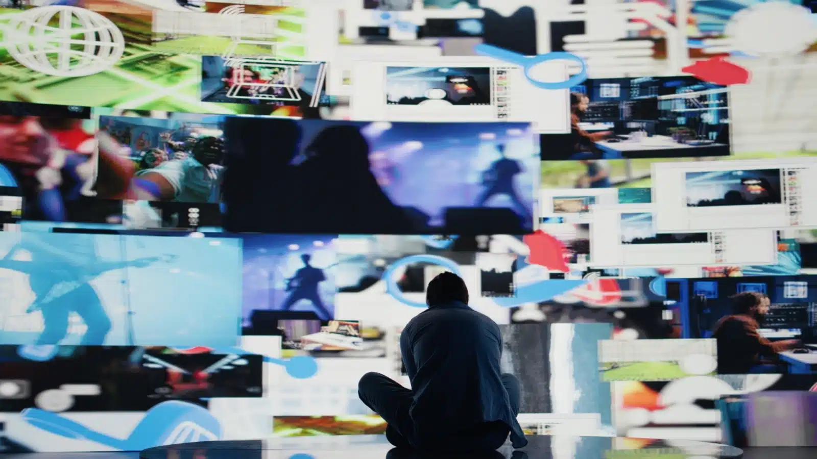 A man sits hunched over in front of a massive screen showing multiple clips at once to represent the things that are wrong with society.