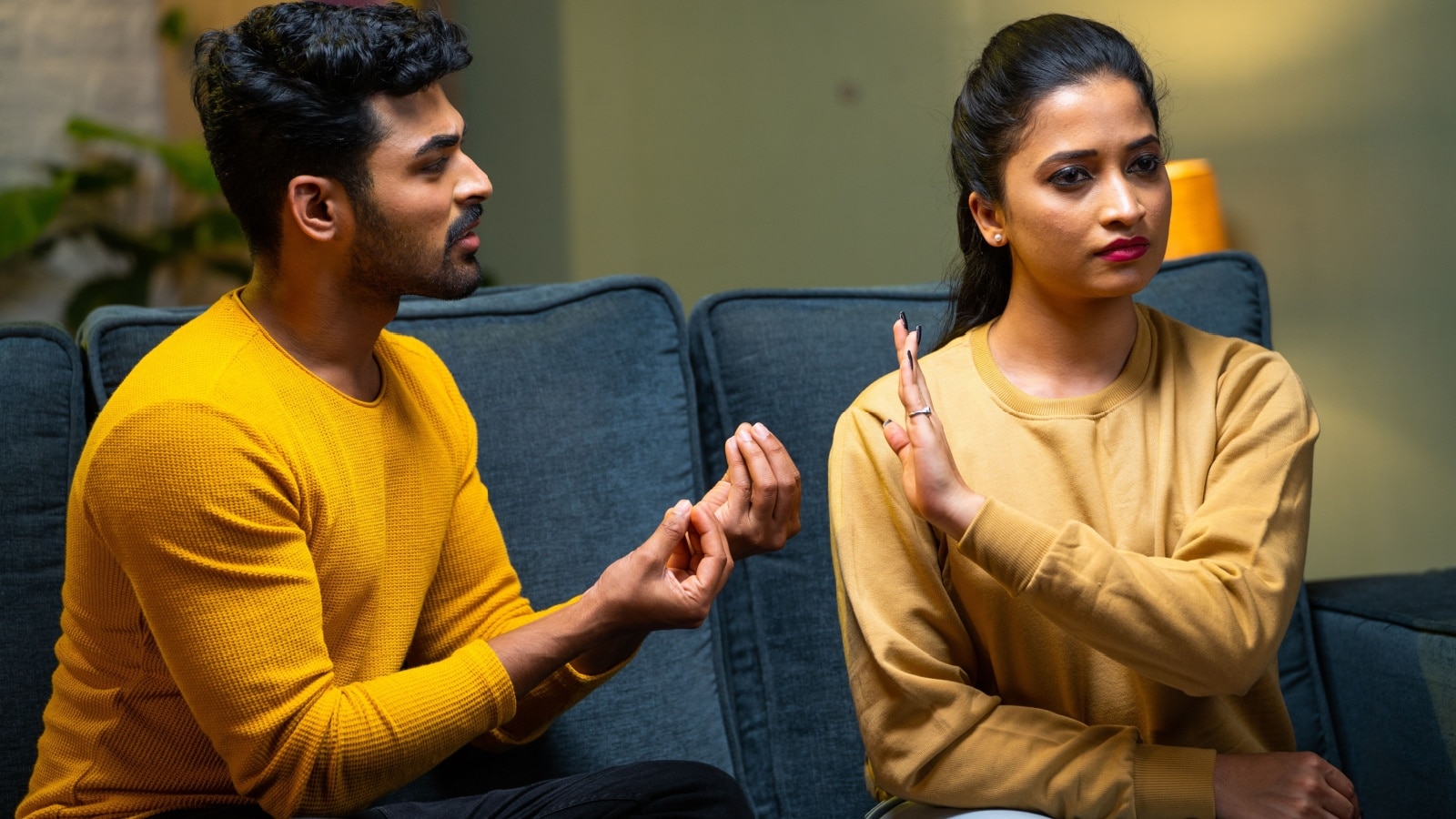 A couple sits on a couch having an argument. She has turned away from him with her hand up, as if she's refusing to listen to his excuses any longer.