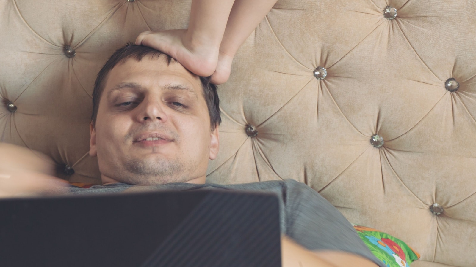 A bad father lays on the bed staring at his screen, ignoring the baby who steps on his head.