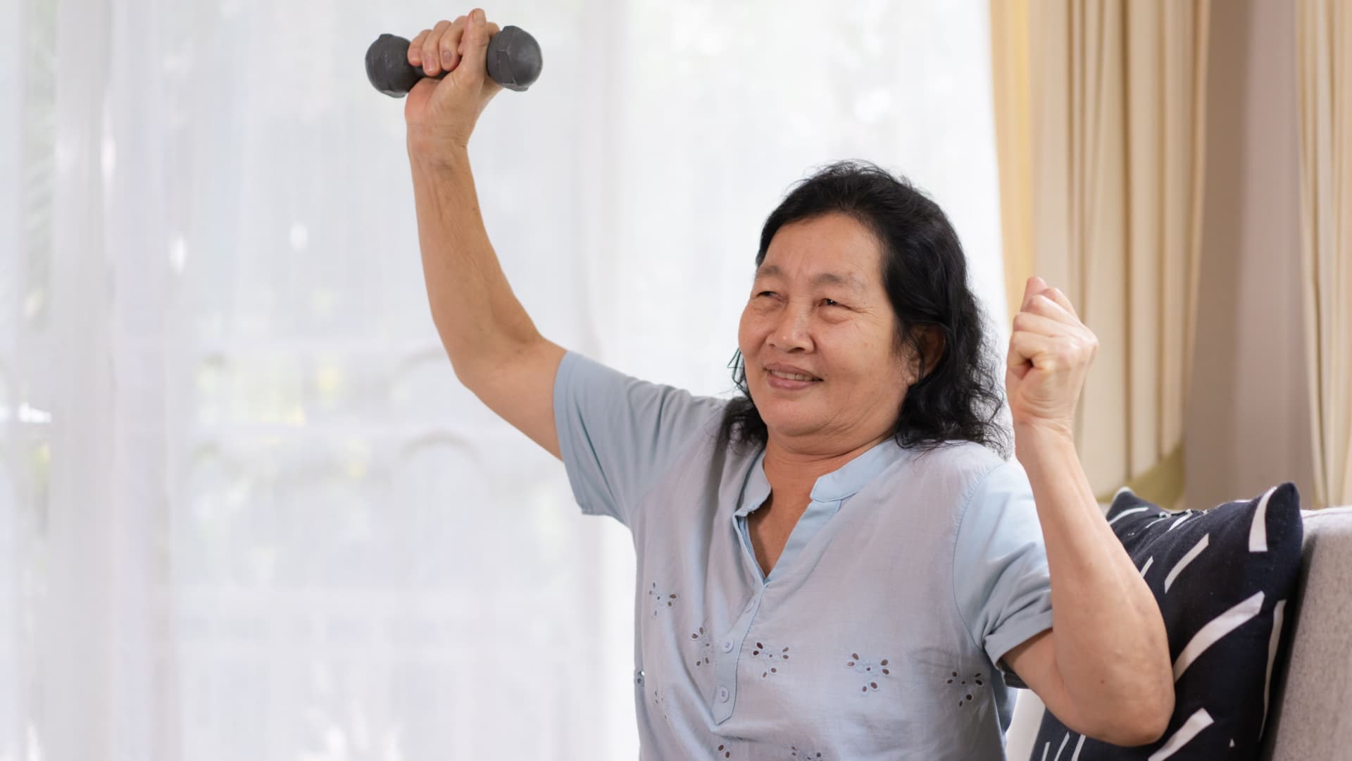 An elderly Asian woman smiles as she lifts a light free weight above her head.