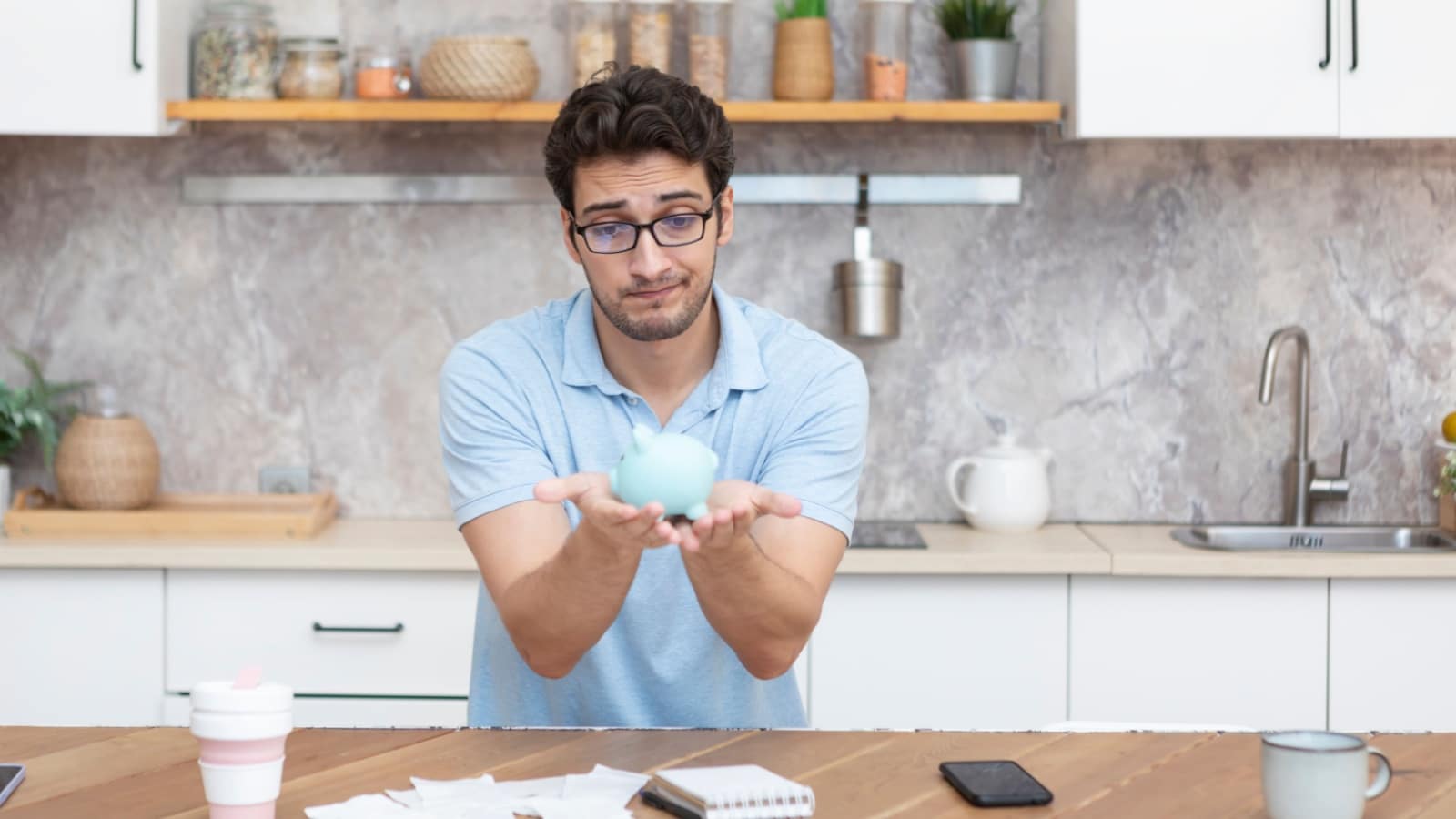 A sad looking man sits at his table with bills in front of him, holding up a tiny piggy bank.