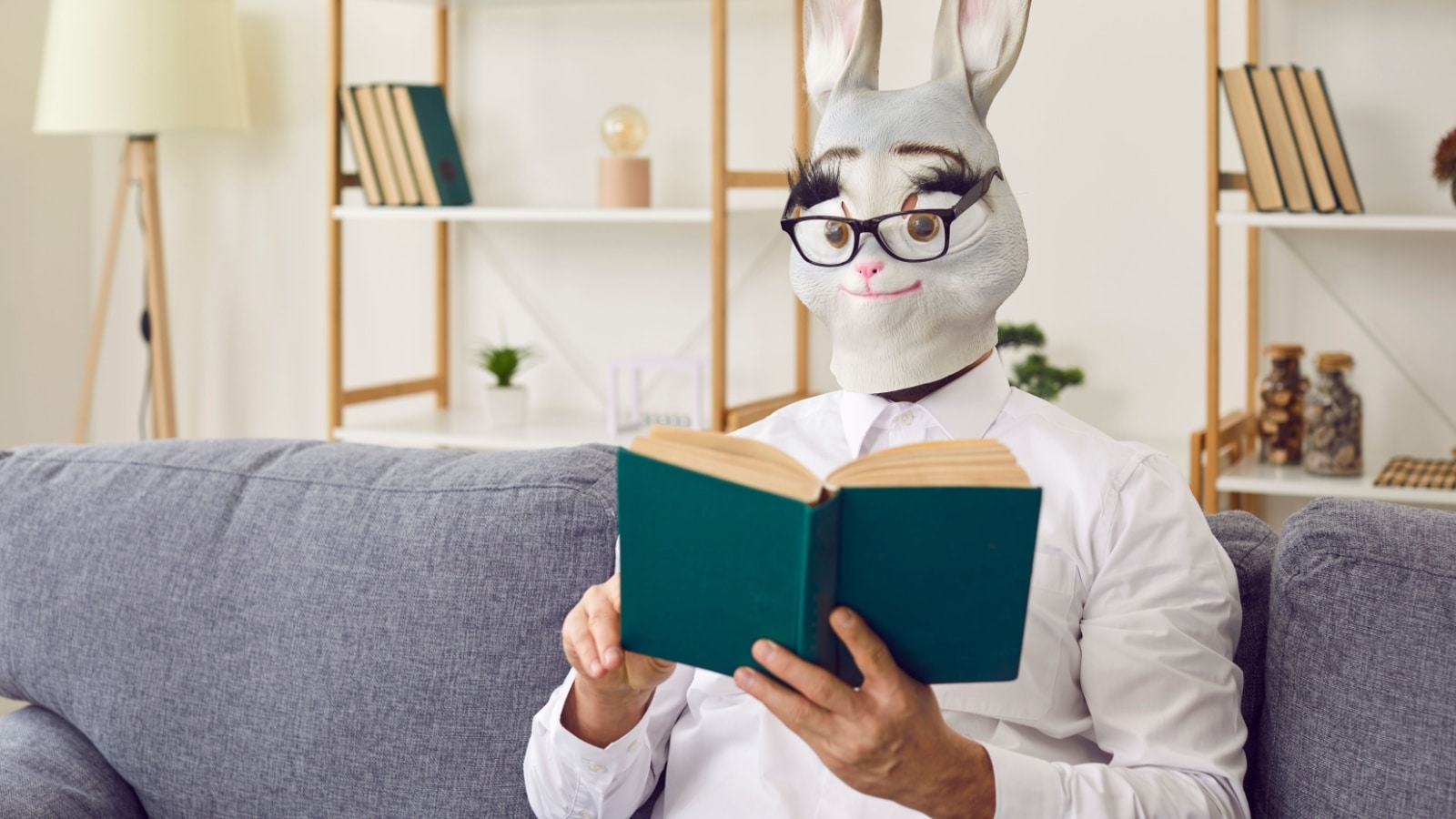 A man wearing a bunny mask sitting on the couch reading a book to represent strange hobbies.