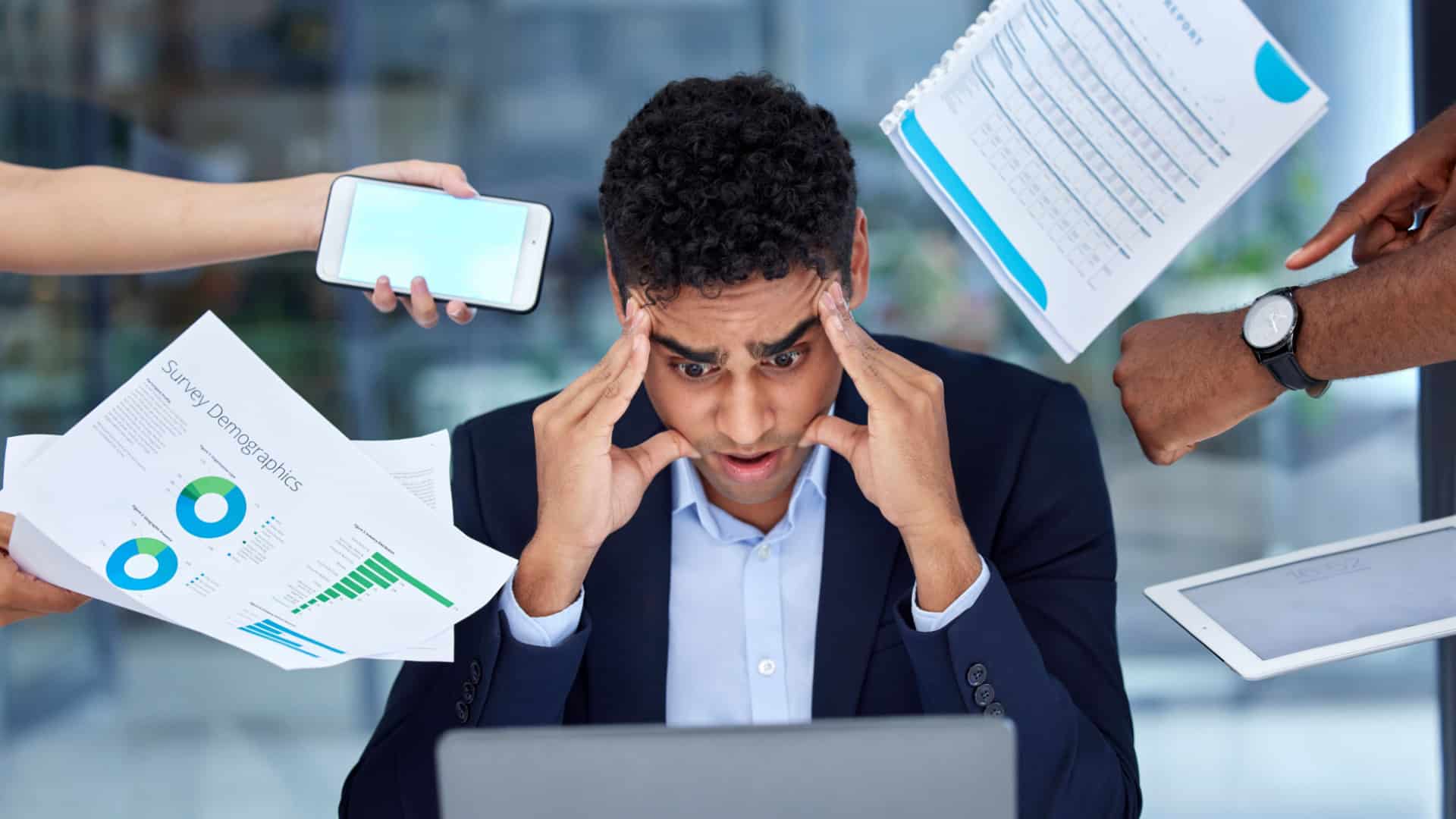 A stressed out worker stares at his laptop screen while hands shove papers and tasks in his face.