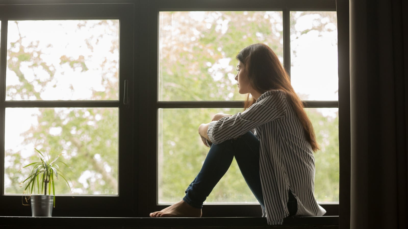 A woman sits on a window sill looking sadly outside to represent this doesn't feel like me.