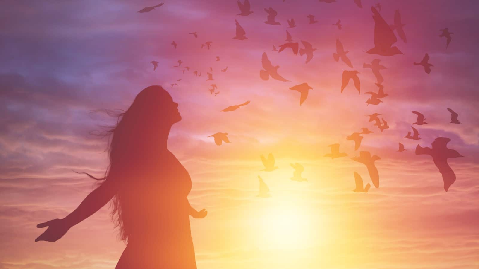 Silhouette of a woman basking in the sun at sunset with birds flying all around her. She has her hands out in a sign of positivity to represent good juju.