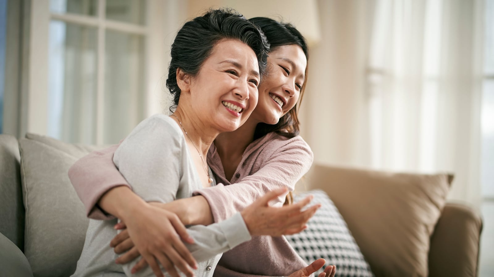 A happy adult daughter hugs her mother on the couch in their home. Both are smiling.
