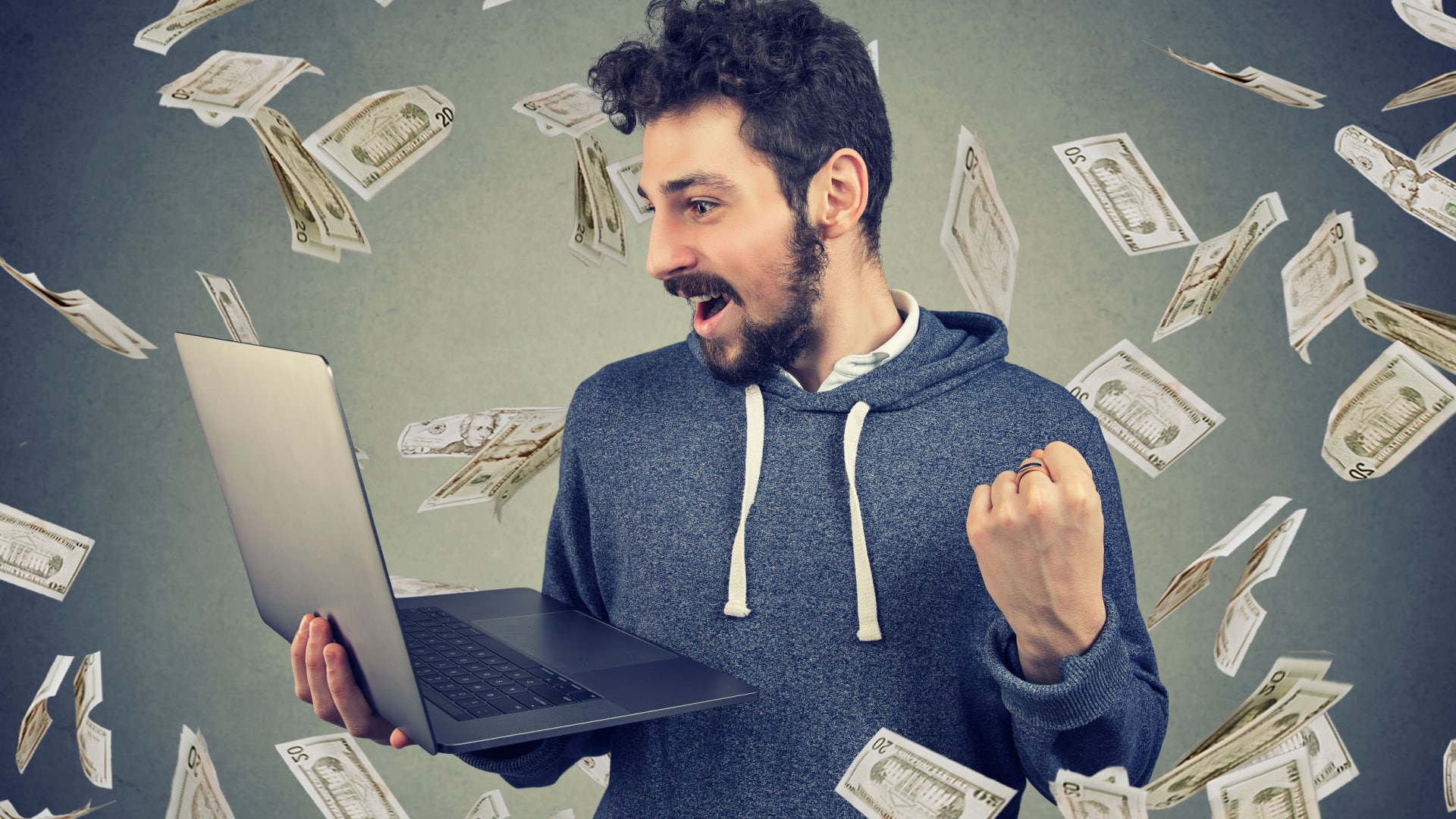 A happy man holds his laptop as money flies around him to show that he's making money online.