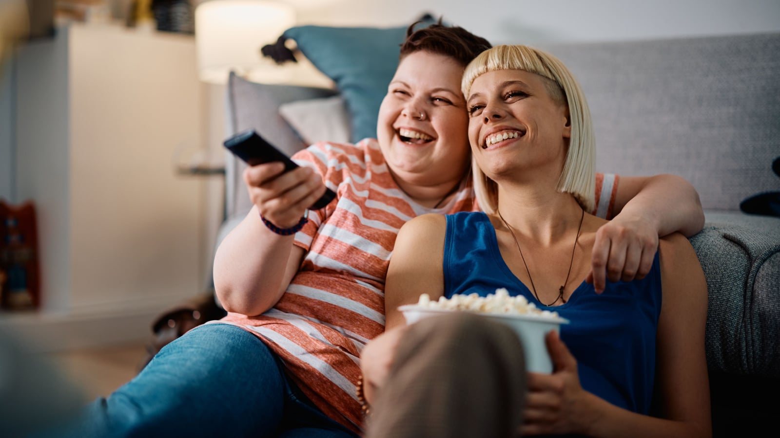 A lesbian couple snuggling on the couch with popcorn watching a movie.