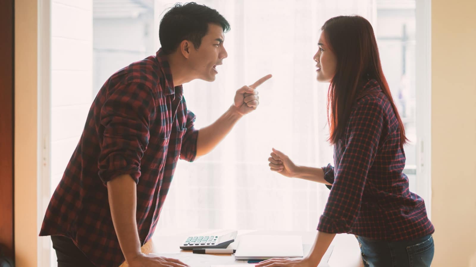 A man angrily pointing his finger and yelling at his wife over a table with a calculator and bills.
