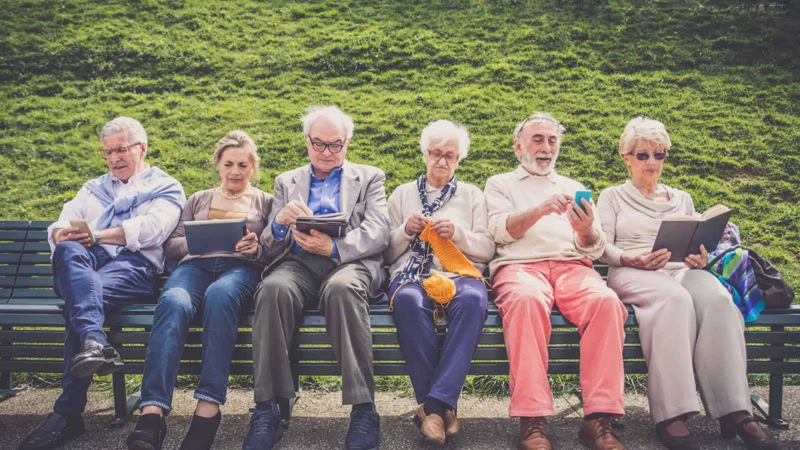 A group of seniors sitting on a park bench. Each is engaged in a different activity, from scrolling on a phone to reading to knitting.