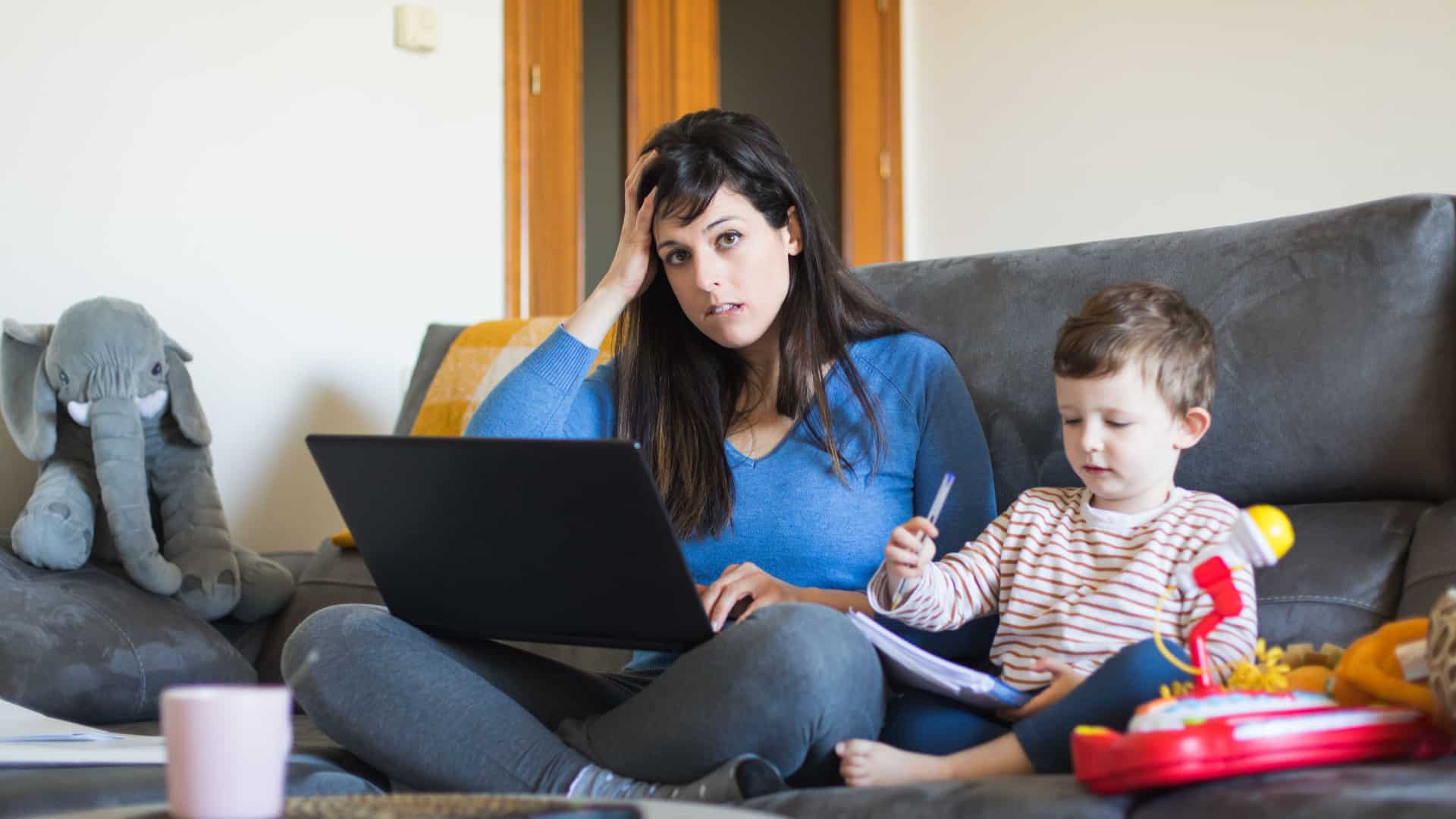 An overwhelmed mom sits on her couch working on a laptop with her young son sitting next to her.