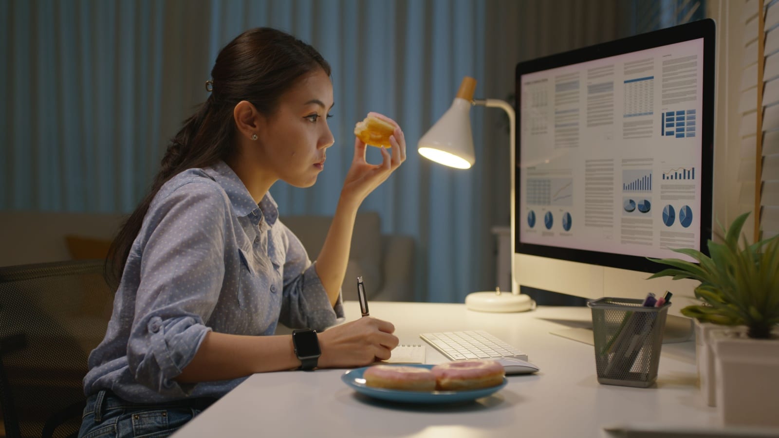A woman sits at her desk at night, working, eating junk food.