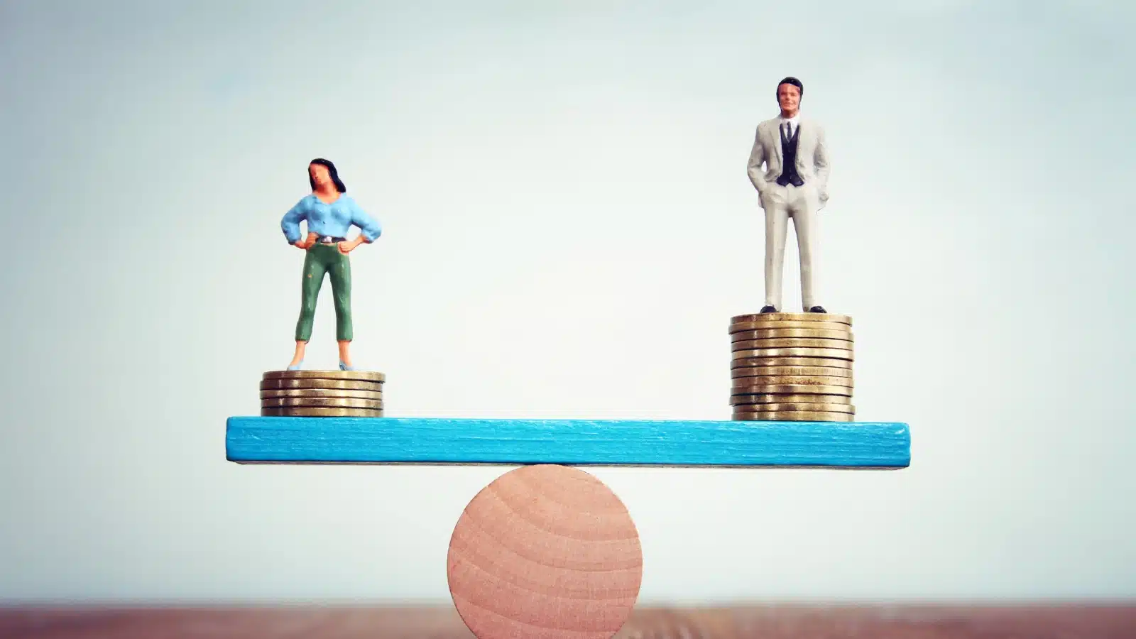A balancing scale represents the gender pay gap. A male figure stands on a higher pile of coins than a female figure, tipping the scale in his favor.