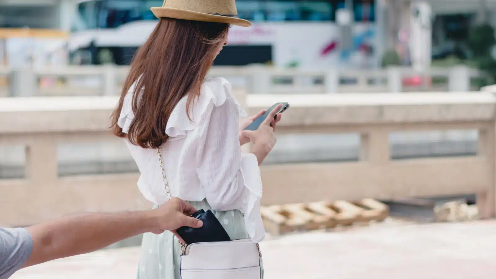 A woman looks at her phone while someone steals her wallet from her purse, to represent how to avoid pickpockets.