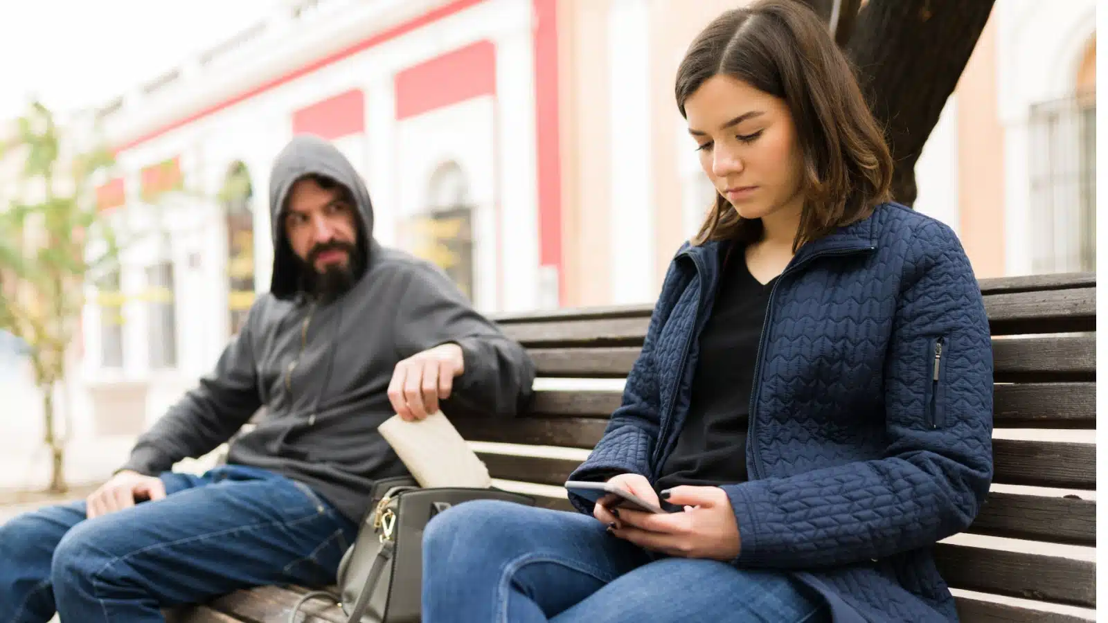 A woman sits on a park bench looking at her phone, oblivious to the man stealing the wallet out of her purse right next to her.