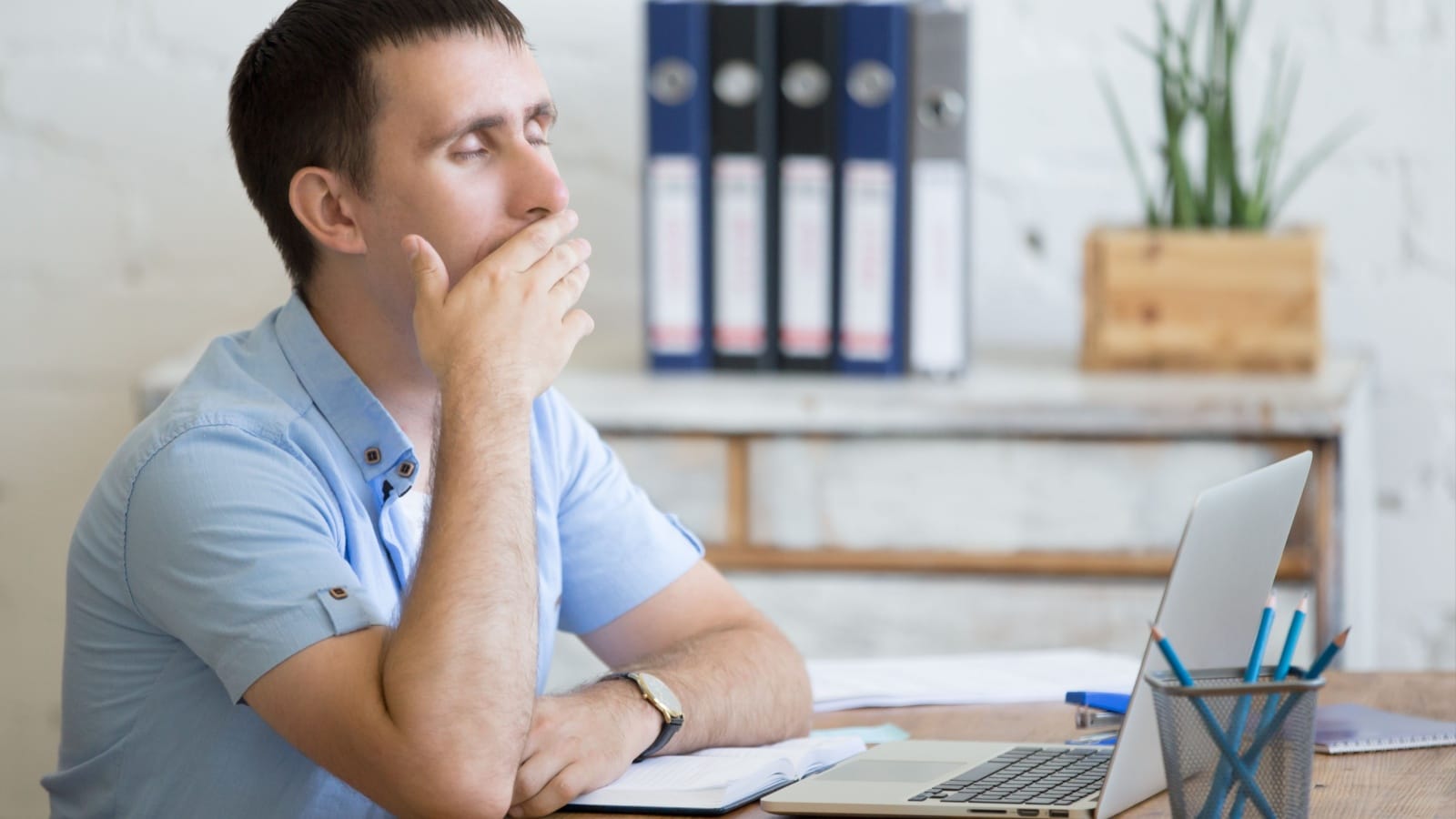 A tired man yawning at his desk in front of his laptop.