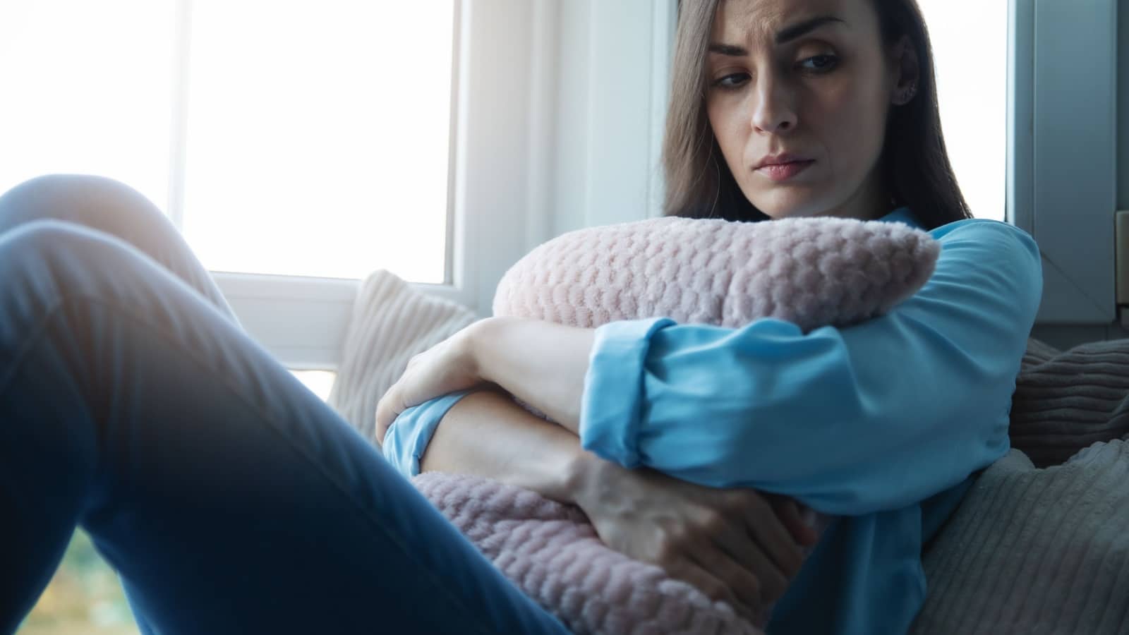 A sad woman hugs a couch cushion as she stares into space, perhaps thinking "what is wrong with me."