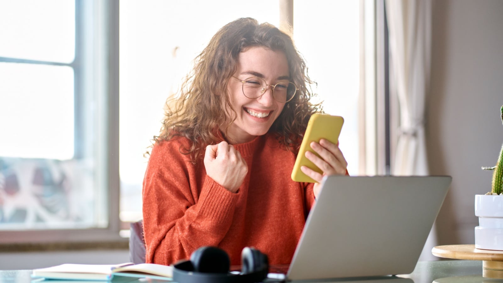 An excited woman looks at her phone while working at a computer as if she just got hired for her dream job.