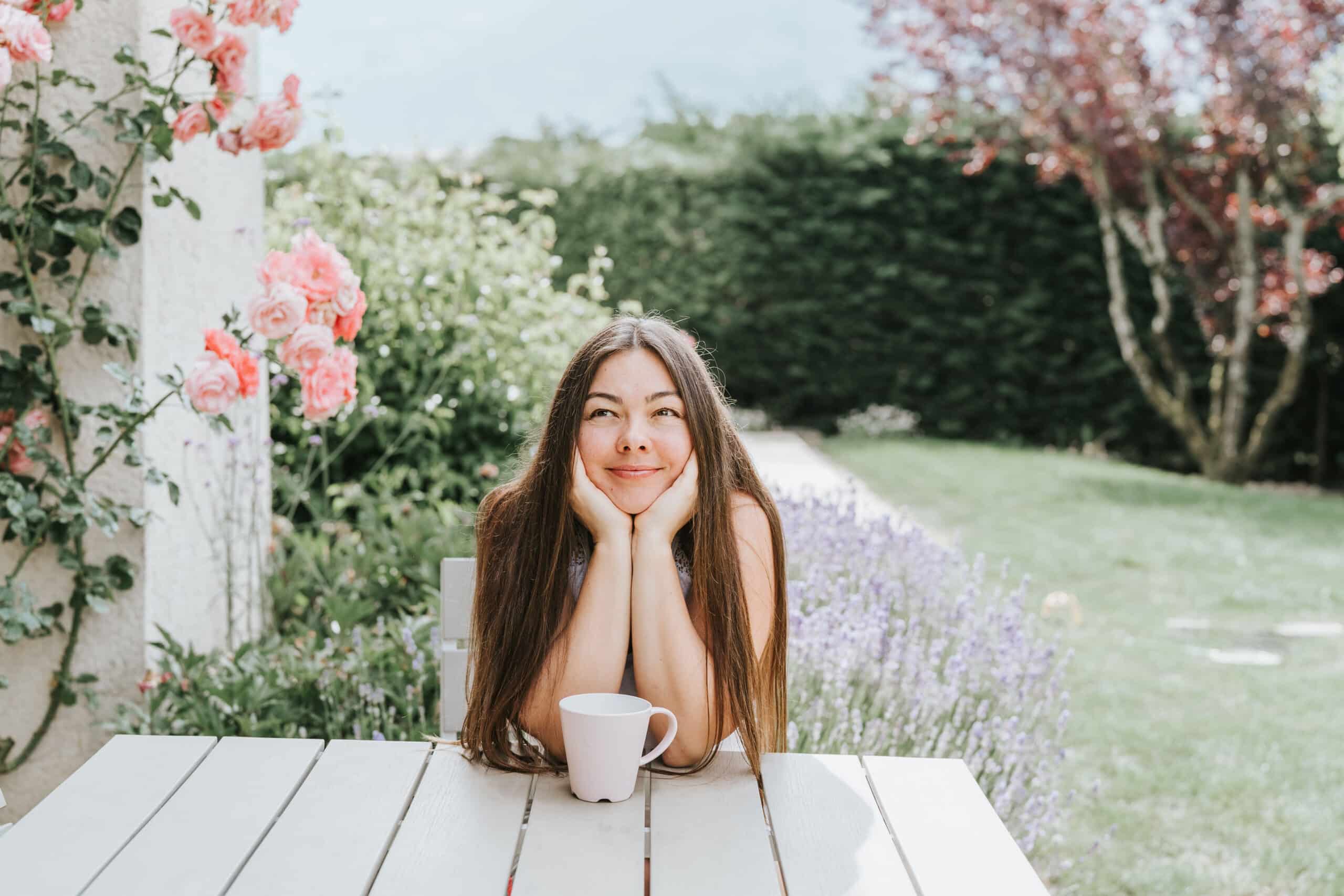 A woman enjoying a cup of coffee outside in the spring. She's sitting at a picnic table in a lovely garden to represent lifes simple pleasures.