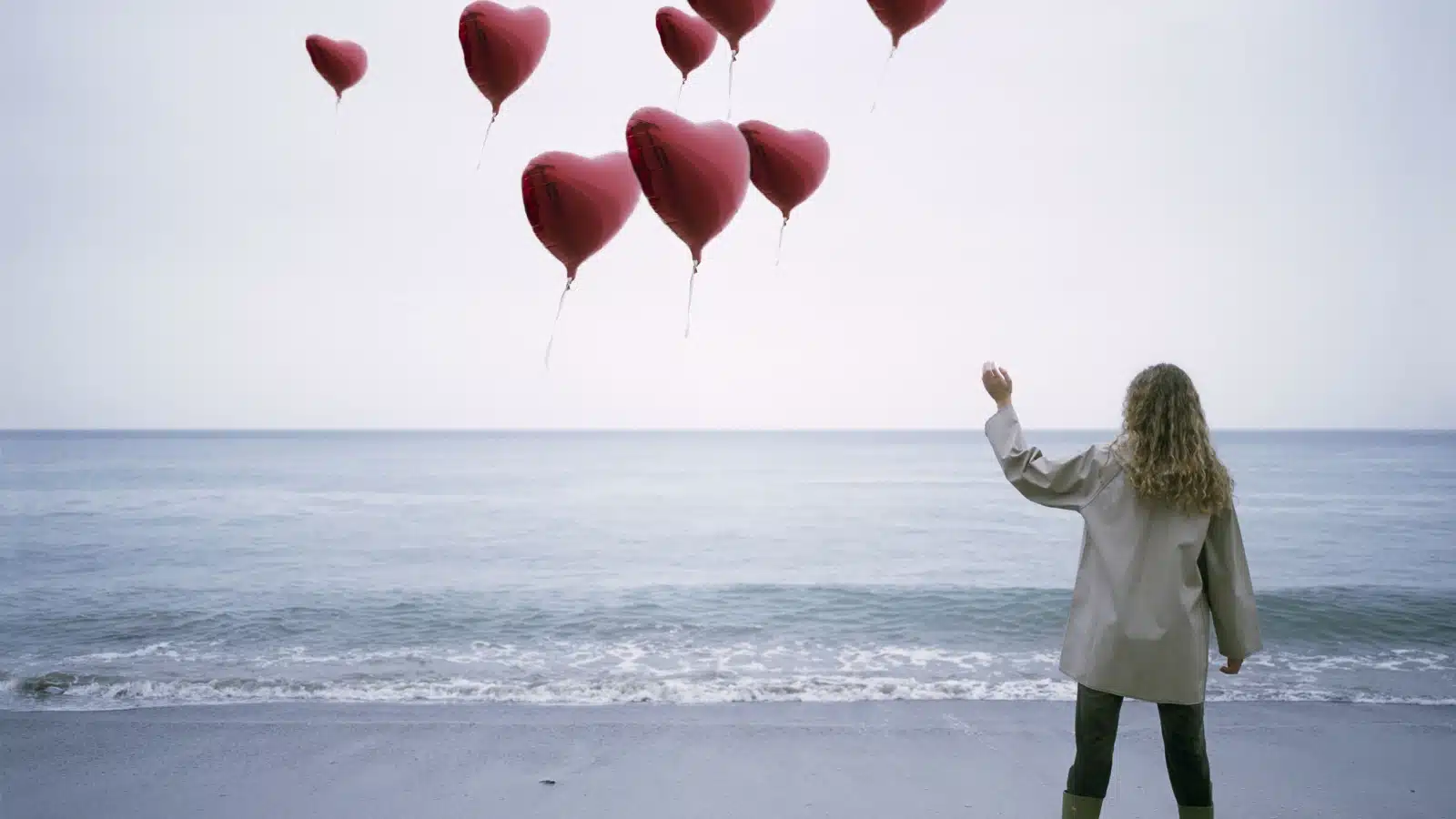 A woman lets go of heart balloons, letting them drift away over the ocean. The image represents acceptance and moving on.