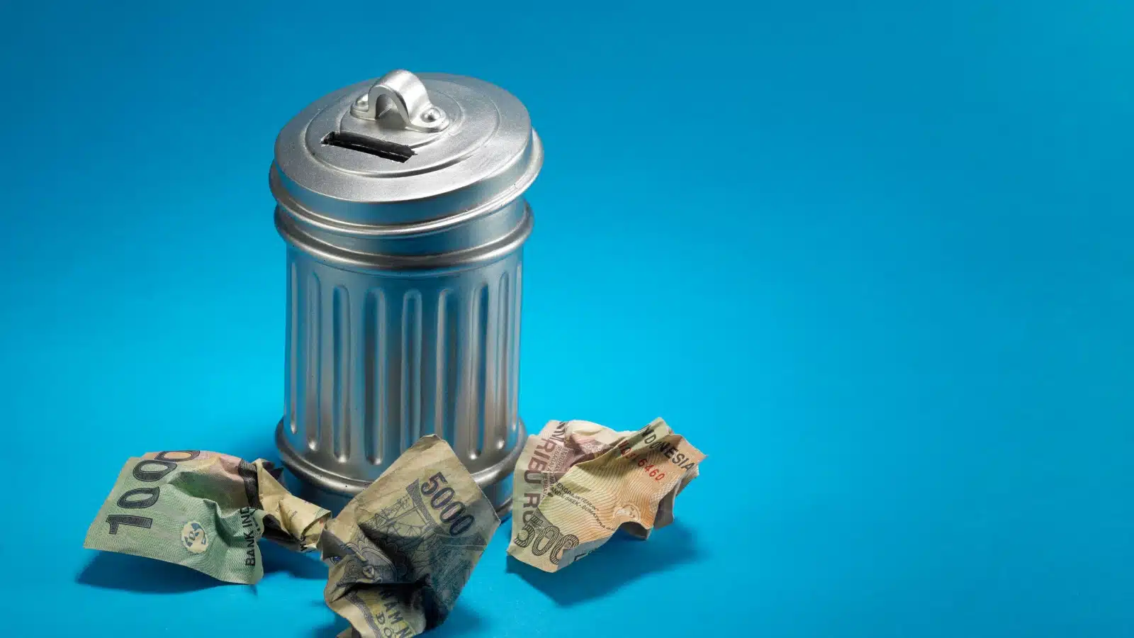 A trash can with crumpled bills around it to represent throwing money away.