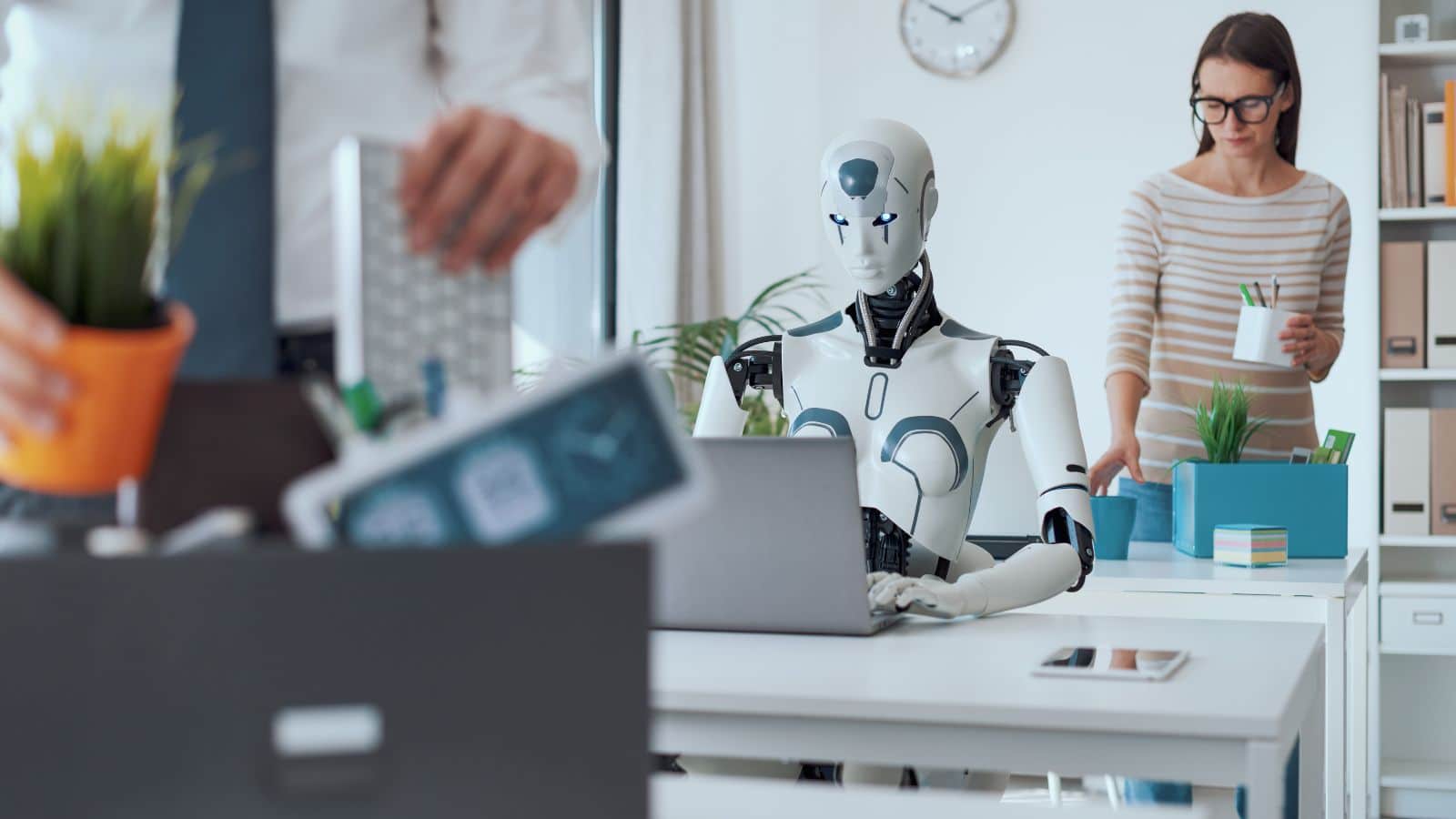 A robot works at a computer, while a blurred human worker in front packs up their desk. The photo showcases how workers are losing jobs to AI.