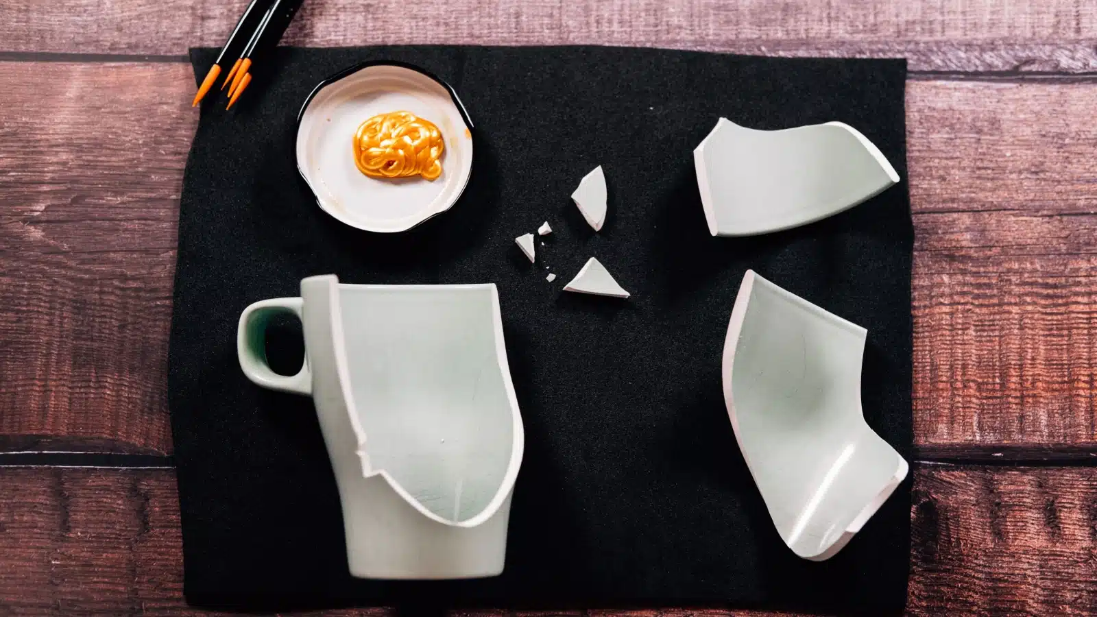 A broken white cup sits on a black velvet cloth with kintsugi paint and brushes, to represent how to get started with Kintsugi.
