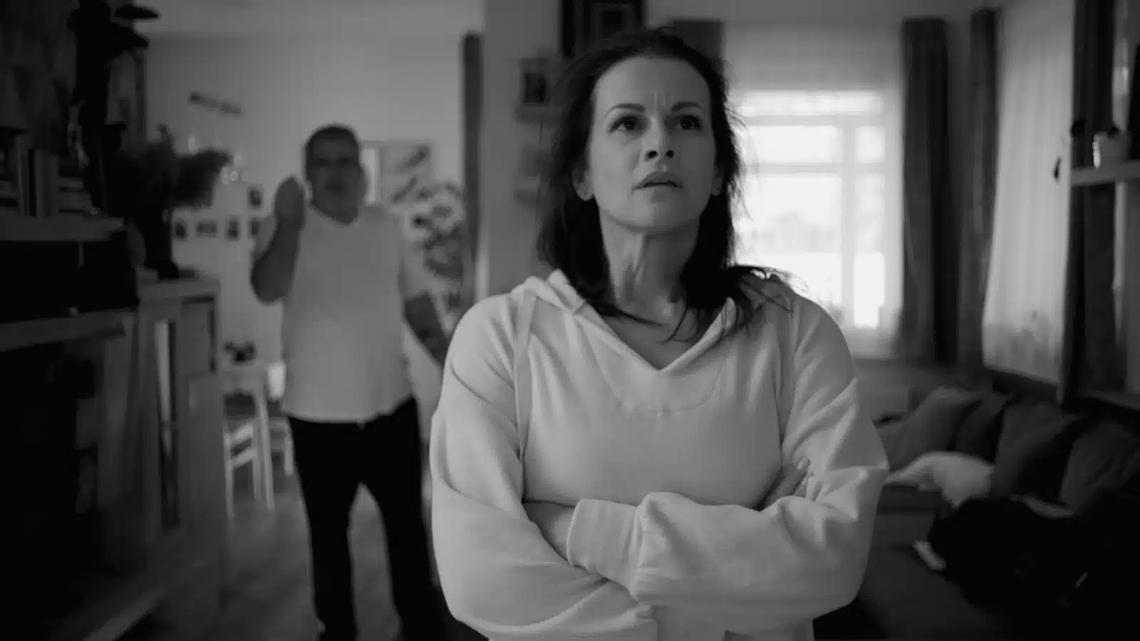 A sad looking woman has her arms crossed while her husband yells at her in the background.