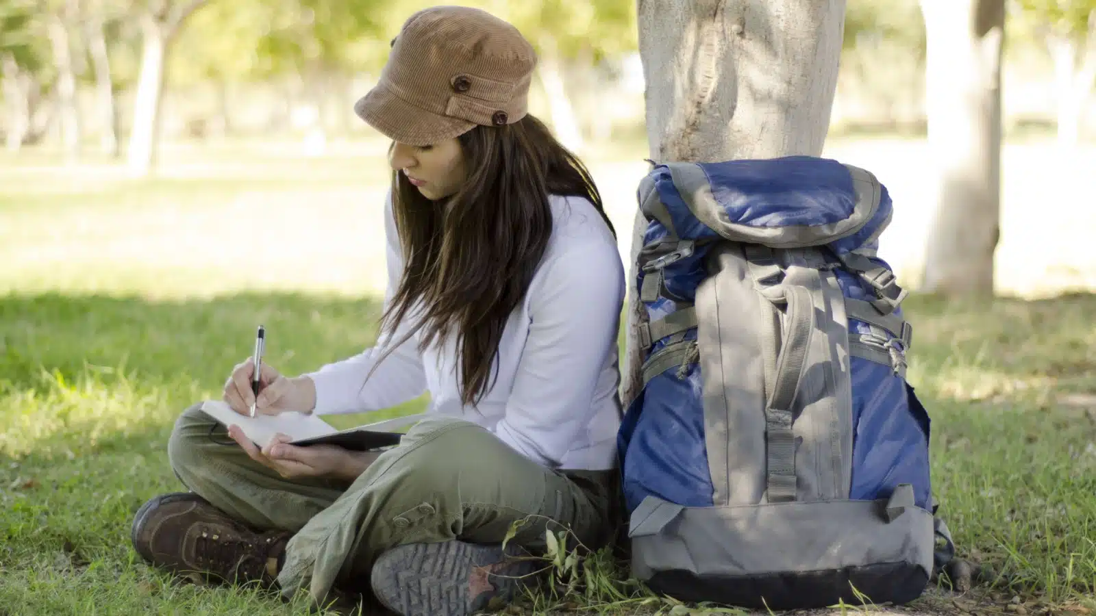 A hiker rests under a tree, writing in her travel journal.