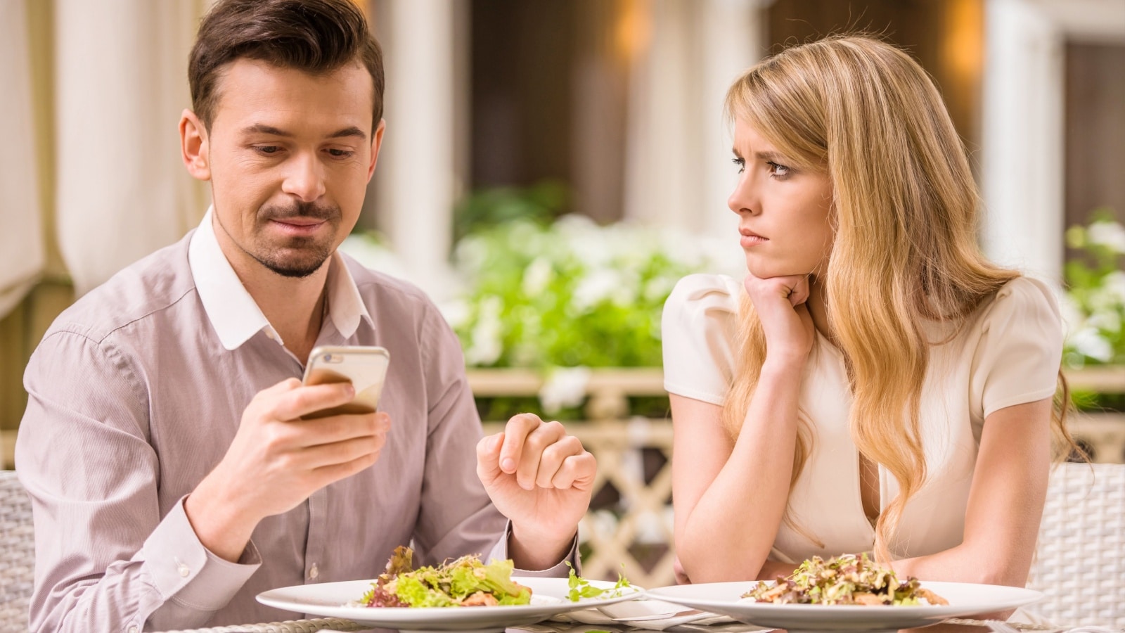He's looking at his phone ignoring his date while at the restaurant. She looks at him as if she's upset.